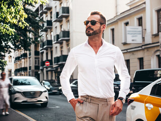 Portrait of handsome confident stylish hipster lambersexual model.Modern man dressed in white shirt and trousers. Fashion male posing in the street background in Europe city at sunset. In sunglasses