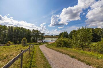 Fototapeta na wymiar Gorgeous views of the natural landscape. Country road to lake between green trees on a blue sky with a background of white clouds. Sweden.