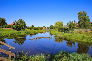 View on quiet peaceful place at dutch hiking trail with idyllic river, lush green meadow and trees against deep blue cloudless summer sky, small village background  - Neer (Limburg), Netherlands
