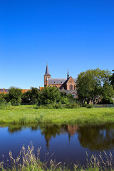 View over river and green pasture on small dutch idyllic village with medieval old church against blue cloudless summer sky - Neer (Limburg), Netherlands