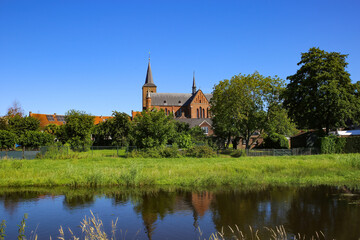 Fototapeta na wymiar View over river and green pasture on small dutch idyllic village with medieval old church against blue cloudless summer sky - Neer (Limburg), Netherlands