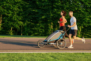 Running couple with child in jogging stroller in public park at sunrise. Morning run for a whole...