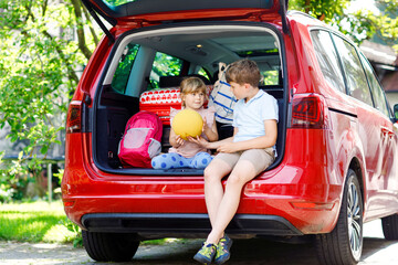 Two children, school boy and preschool girl sitting in car trunk before leaving for summer vacation with parents. Happy kids, siblings, brother and sister with suitcases and toys going on journey