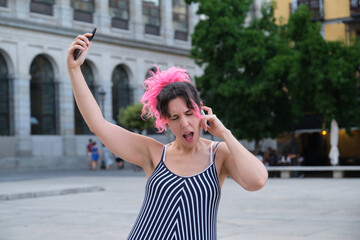 Young caucasian woman with pink hair dancing in street with earbuds and phone.