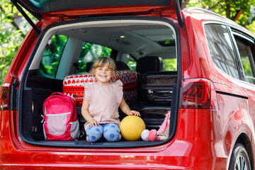Cute little preschool girl sitting in car trunk before leaving for summer vacation with parents. Happy active child with suitcases, bike and toys going on family journey, road trip