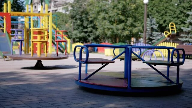 Rotation of an Empty Multicolored Carousel on a Playground in a City Park. Deserted park in summertime. People, children stay at home on self-isolation. Pandemic, quarantine, covid-19. Slow-motion.