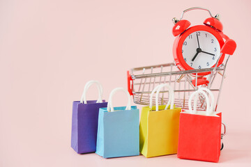 multi color shopping bags , shopping cart and blurred red alarm clock on pink background