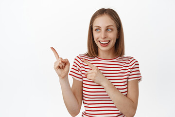 Cheerful young caucasian woman with blond hair, laughing and smiling happy, pointing fingers left, looking at sale promo text, showing advertisement, white background