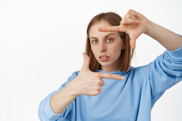 Image of serious creative blond woman searching angle for shot, look through hand frames with furrowed eyebrows, thinking, working on perfect photo, measure something, white background