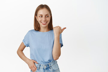Cheerful blond woman smiling white teeth, pointing right with happy face, checking out advertisement, showing promo text, standing in t-shirt against white background