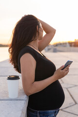 Pregnant hispanic woman holds smartphone and relax on embankment with warm light in morning