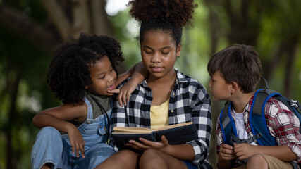 Sister black skin African American ethnicity sitting at on tree base nature reading to her younger siblings a book about adventure stories