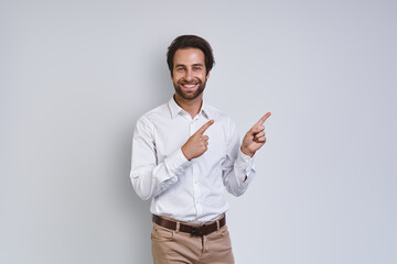 Handsome young smiling man in white shirt looking at camera and pointing away while standing against gray background