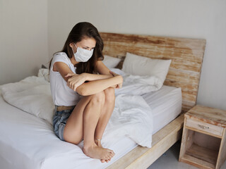 woman in a medical mask in shorts and a t-shirt sits on the bed on vacation in quarantine coronavirus