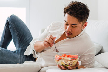 Portrait of young handsome Chinese man eating a fresh, delicious and healthy tomate salad with tune on the sofa in the living room of a minimalist and clean home