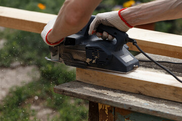 A carpenter planks wood with an electric plane. Woodworking carpentry. Close-up. Man in the garden sanding wooden planks. DIY home improvement, restoration, carpentry concept. Sawdust flies