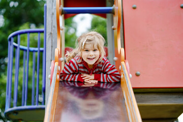 Little preschool girl playing on outdoor playground. Happy toddler child climbing and having fun with summer outdoors activity. Girl slinding down the slide.