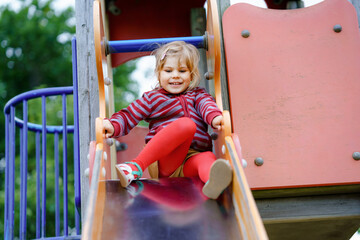 Little preschool girl playing on outdoor playground. Happy toddler child climbing and having fun with summer outdoors activity. Girl slinding down the slide.