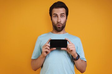 Surprised young man in casual clothing showing his smart phone and smiling while standing against yellow background