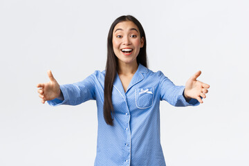Friendly asian girl in blue pajamas welcome girlfriends to her sleepover party, smiling happy and...