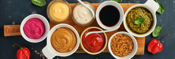 Set of sauces in bowls - ketchup, mayonnaise, mustard, soy sauce, bbq sauce, pesto, chimichurri, mustard grains on dark stone background. Top view copy space.