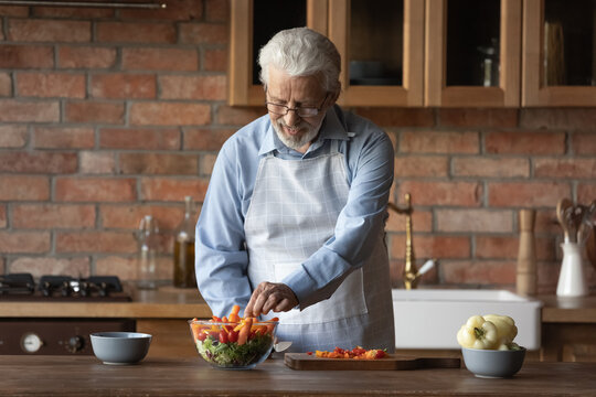 Happy middle-aged 60s Caucasian man in apron stand at kitchen counter prepare healthy tasty vegetable salad for lunch dinner. Smiling old male cook delicious food meal at home. Hobby concept.