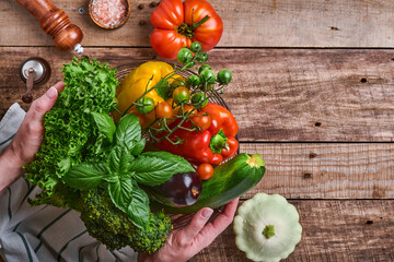 Female hand holding a basket with fresh vegetables and fruits on old wooden rustic background, top view. Space for text. Food cooking background and mock up.