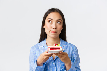 Close-up shot of thoughtful cute asian girl standing at night in pajama with plate of cake, looking...