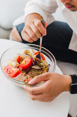Close-up of young handsome Chinese man eating a fresh, delicious and healthy tomate salad with tune on the sofa in the living room of a minimalist and clean home