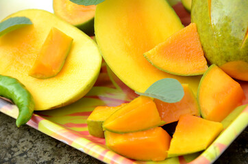 closeup the sliced ripe green yellow mango fruit with green mint leaves and chilly in the tray over out of focus grey brown background.