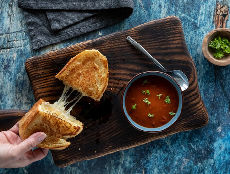 Hand taking one half of a grilled cheese sandwich and a bowl of tomato soup. Soup and sandwich combo concept.