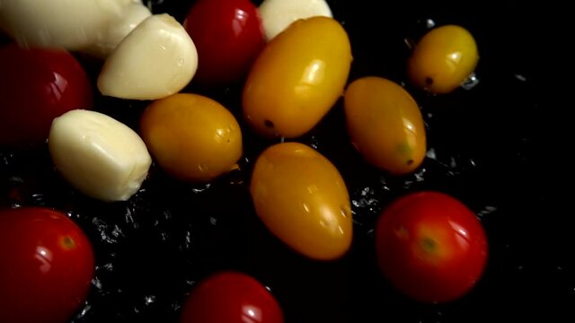 Falling red and yellow cherry tomato and garlic into water. Washing tomatoes. On a black background. Slow motion.