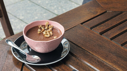 A pink bowl with chocolate mousse, pieces of walnut and a teaspoon on the dark brown table at cafe