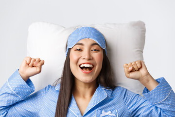 Close-up of enthusiastic asian girl in blue pajamas and sleeping mask, stretching hands up delighted after good night sleep, take-off eyemask in morning, lying in bed on pillow and smiling happy