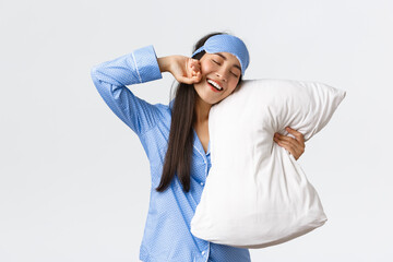 Smiling pleased cute asian girl in blue pyjama and sleeping mask, hugging pillow and stretching...