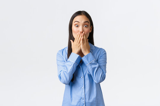 Shocked Startled Girl Witness Something Horrible And Surprising. Asian Woman In Blue Pajamas React Astounded, Cover Mouth With Hand In Awe And Stare Camera Speechless Over White Background