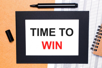 On a wooden table, there is a black open marker, a brown notepad and a sheet of paper in a black frame with the text TIME TO WIN. View from above.