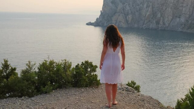 A woman in a white dress is walking in the mountains near the seashore, enjoying a beautiful view of the bay