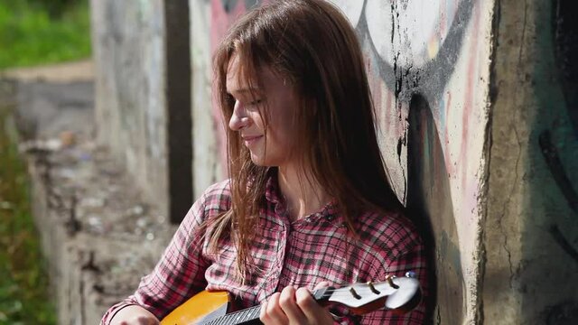 A beautiful emotional girl with long hair plays a musical instrument against the background of a concrete wall. Strong wind blows the hair. Domra