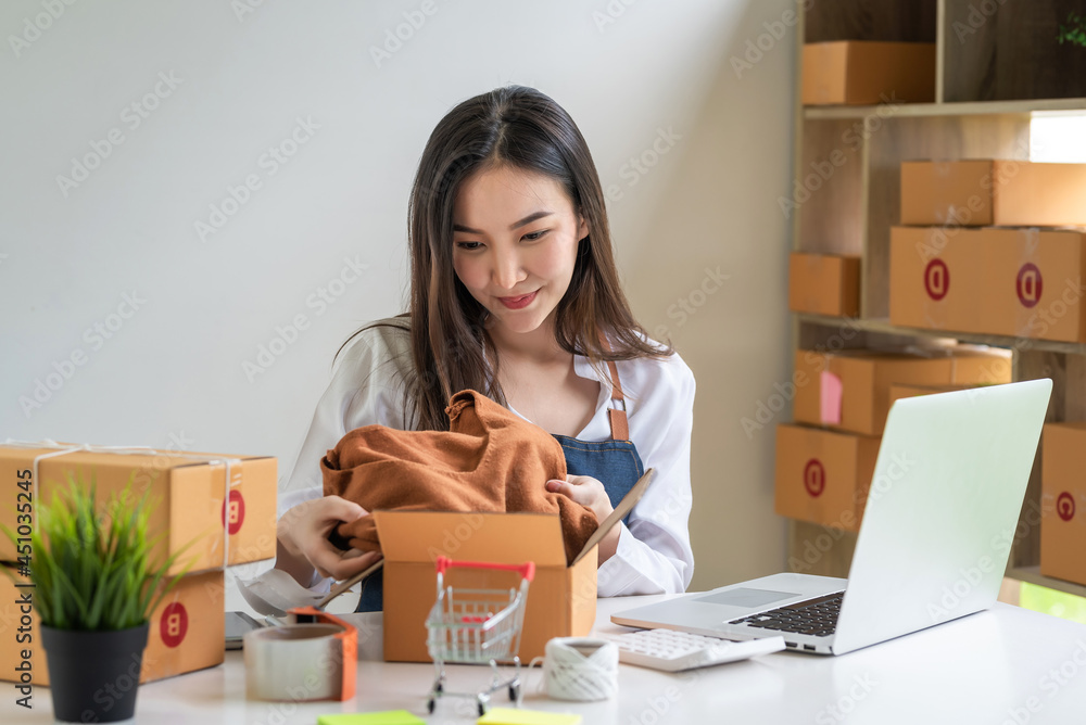 Wall mural Business owner small online woman is putting items in a parcel box to deliver to customers calculator laptop on the desk.