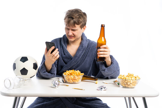 cheerleader, a man in a robe sits at a table and watches TV. empty bottles and wrappers are scattered across the table. a man is a fan of his favorite team, drinks beer