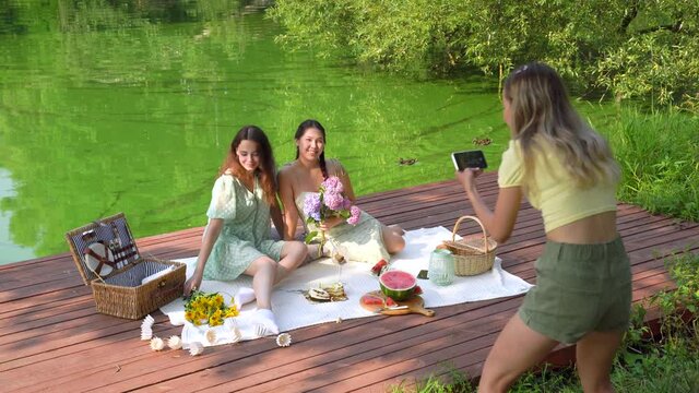 blond young female is taking photo of her asian friends on picnic dinner.  Attractive girls having lunch on nature. High-angle shot