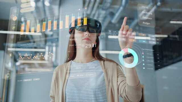Young Adult Female Using Futuristic Augmented Reality Software Interface for Managing Business and Marketing Projects. Specialist in Office Wearing Headset to Look at VFX Animation with Financial Data