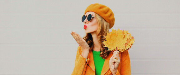 Autumn portrait of beautiful young woman with yellow maple leaves blowing her lips with red lipstick wearing a french beret on gray background