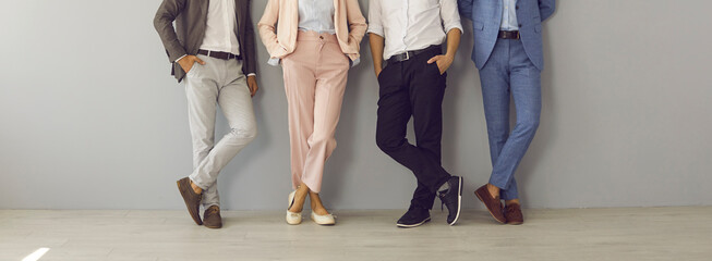 Group of four successful company leaders in classy formal suits standing hand in pocket. Team of 4 business partners leaning on grey office wall. Cropped shot of people's legs in stylish classic pants