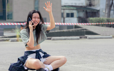 Beautiful happy Asian extreme hipster woman smiling, talking mobile phone and waving hand for greeting, sitting on skateboard, relax playing outdoor activity on holidays in summer vacation.