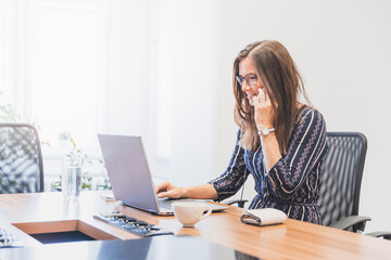 Business woman is sitting at her desk, working on her laptop and talking on the phone, modern workspace, office interior