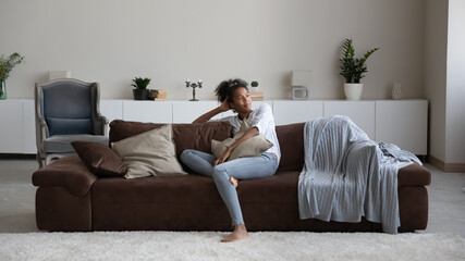 Happy calm millennial African American woman sit relax on sofa in living room look in distance dreaming. Smiling young mixed race female rest on couch at home breathe fresh ventilated condition air.
