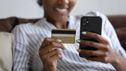 Crop close up of African American woman shopping online at home on cellphone with debit credit card. Smiling ethnic female buyer pay buy on internet on smartphone use secure banking system.