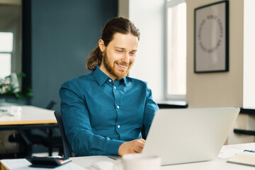 Happy smiling young male office employee in shirt sitting at desk in front of laptop computer and looking at screen with smile, enjoying work in office. Cheerful man using digital technologies at work